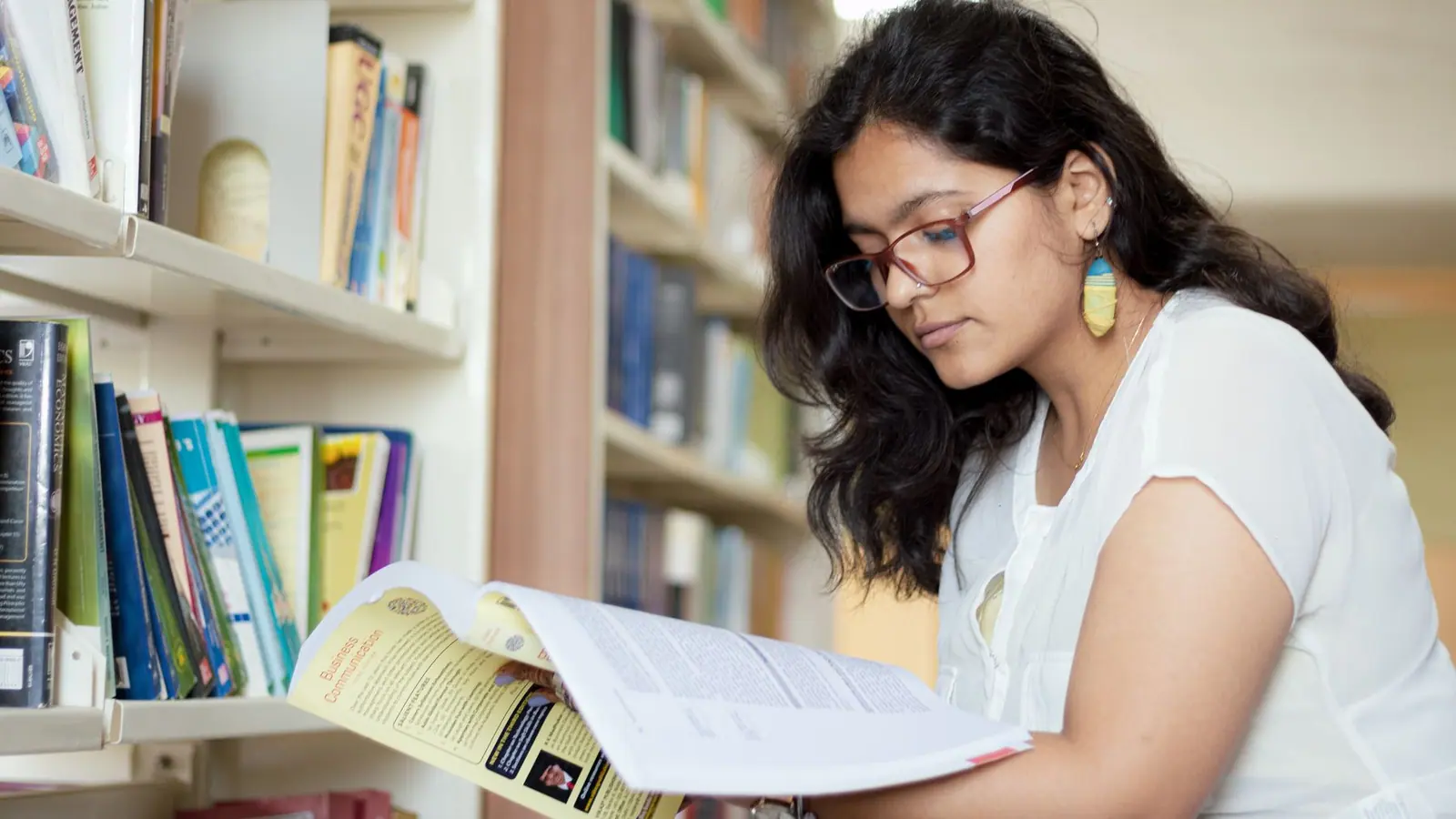 A student reading a textbook in a library beside shelves of books