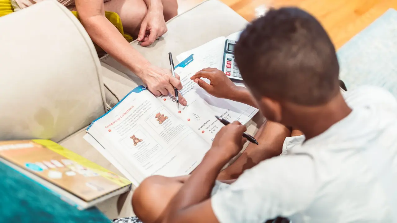 An adult helping a boy with a mathematics workbook at home, with a calculator and pens on the sofa. Stock photograph, not an Infinity Learning student or tutor.